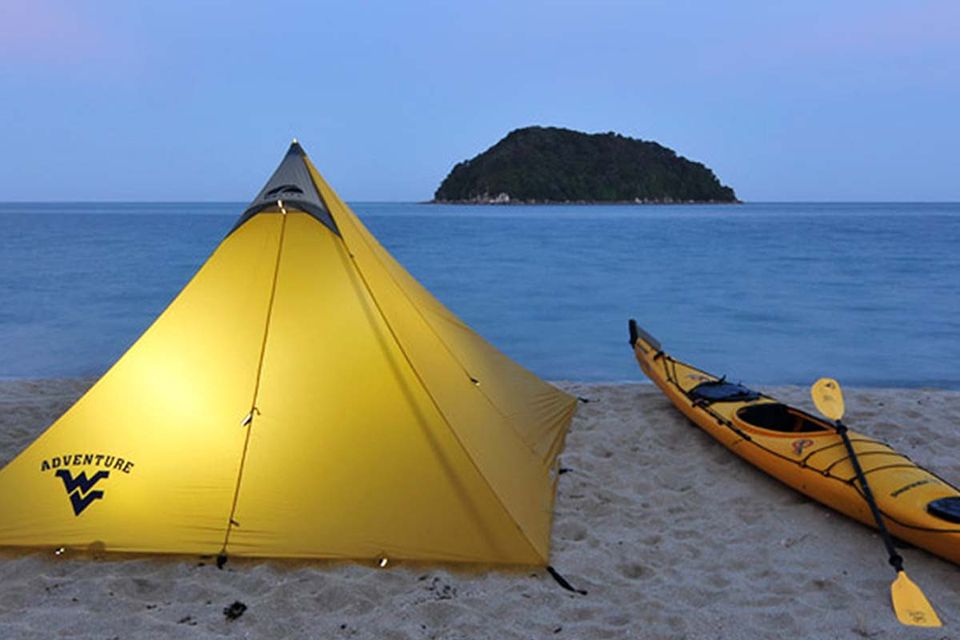 A yellow tent lit from within and a canvas to the left sitting on a beach at dusk in New Zealand.