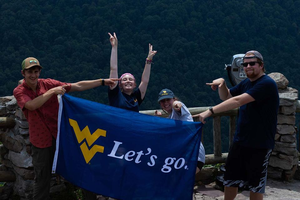 Four excited people at the Coopers Rock Outlook holding a a blue flag with the WVU logo that reads "Lets go.".