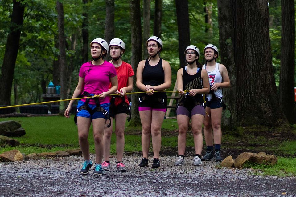 Five students in helmets standing in a line holding onto the same rope in the forest.